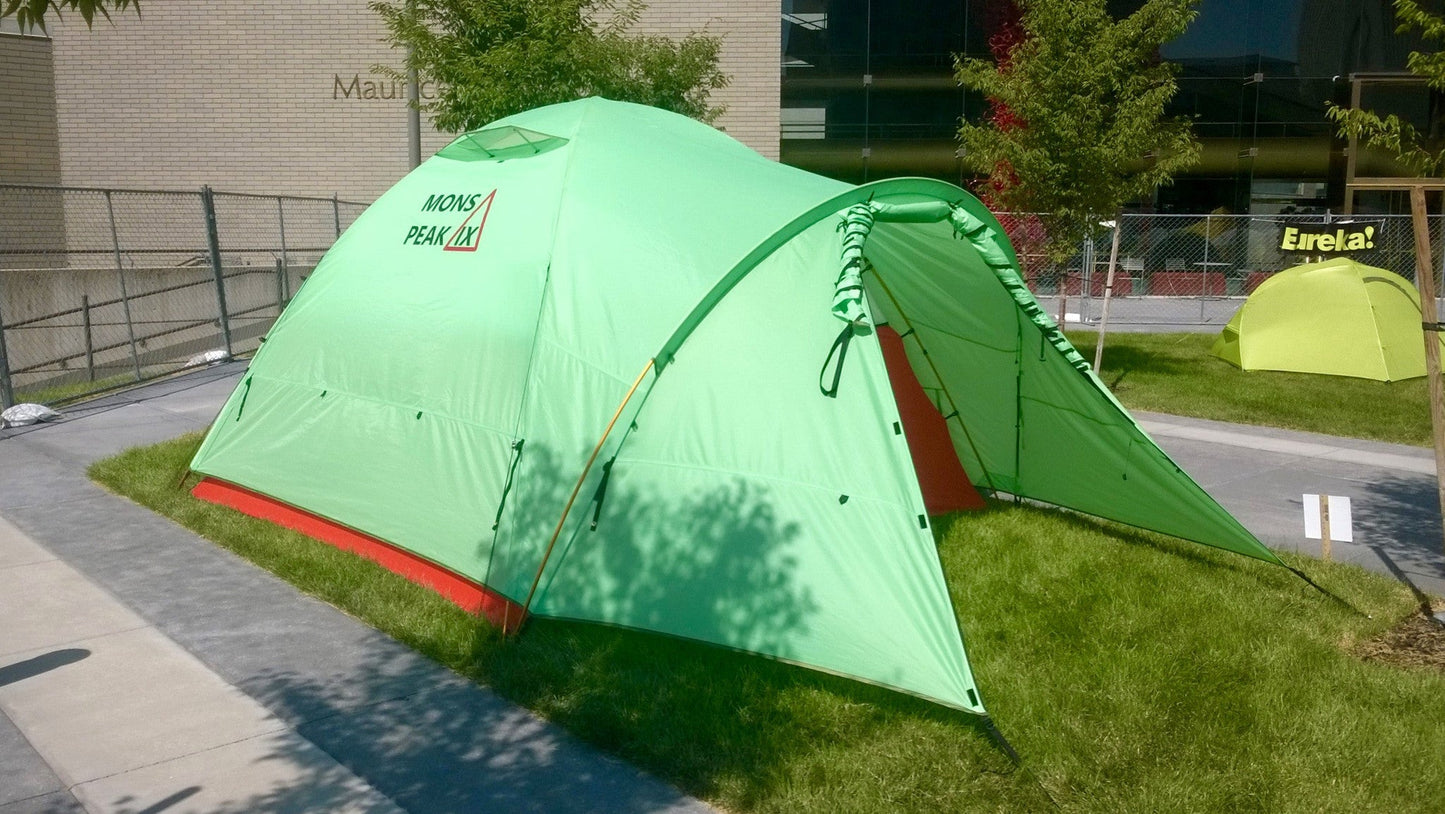 Bright green Mons Peak IX 3-season dome camping tent pitched in urban outdoor setting showing spacious design, red groundsheet, large front vestibule, and Mons Peak IX logo on fly