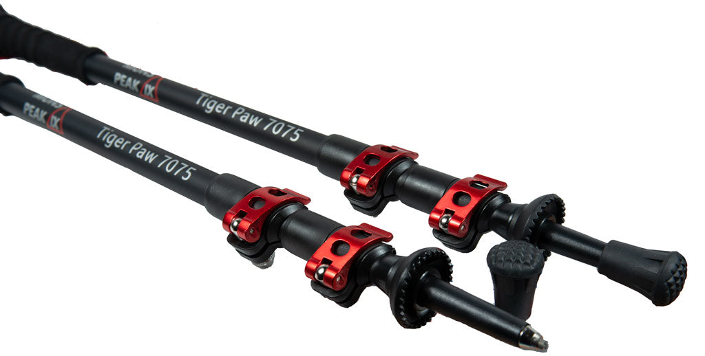 Close-up of Mons Peak IX Tiger Paw 7075 carbon trekking poles showing black shafts with branding, red quick-lock collars, foam extensions, oversized mud/snow baskets, and carbide spike tips